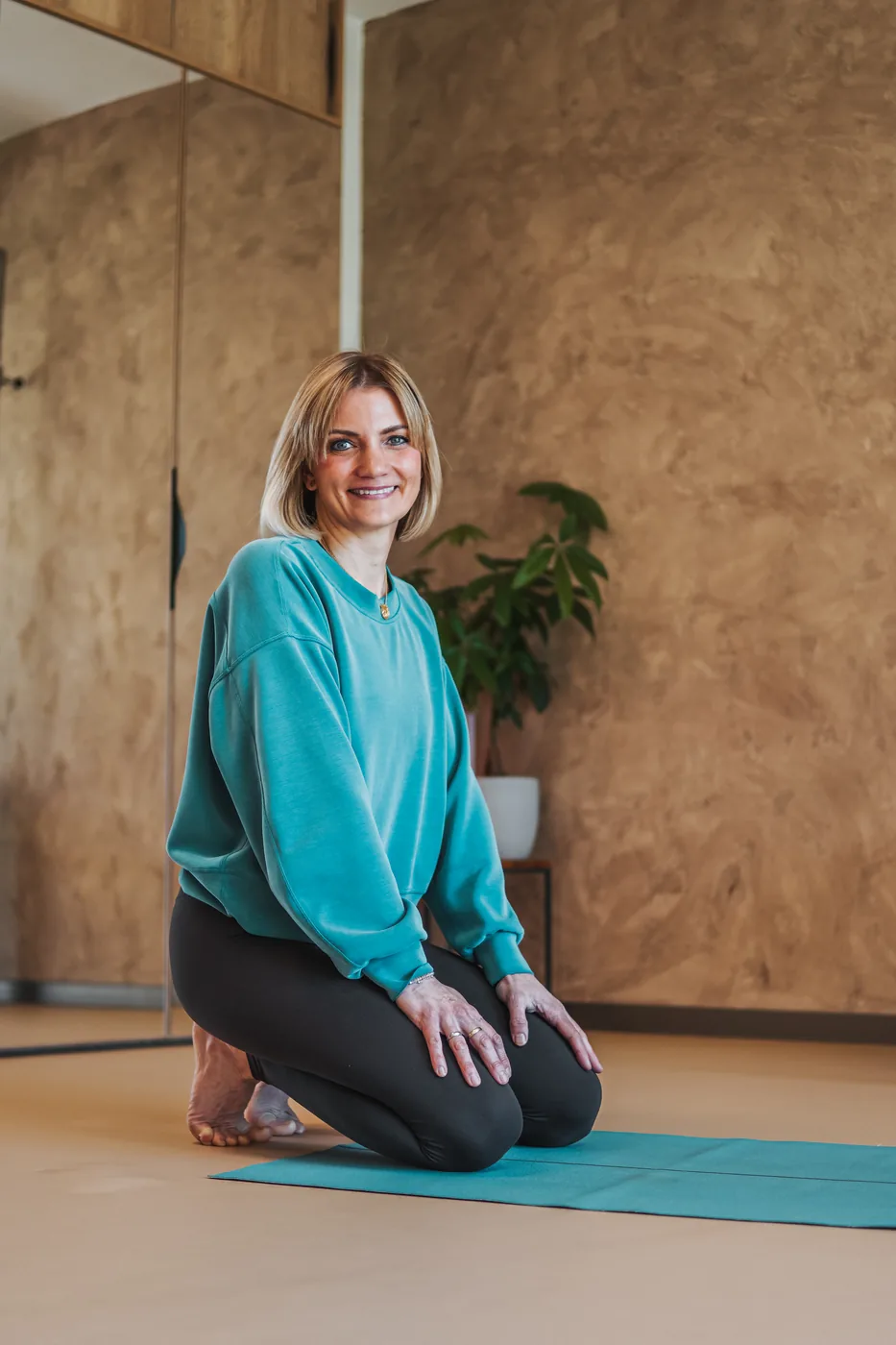 Lenka Vokřínková klečí na podložce ve studiu s hliněnou stěnou a usmívá se